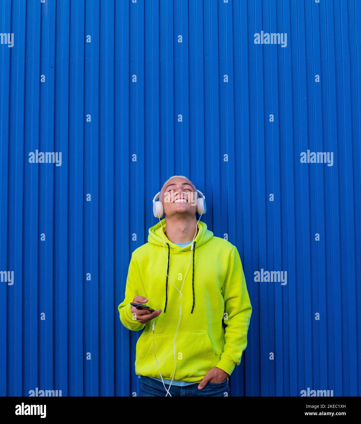 un joven o adolescente milenario escuchando música con su teléfono y auriculares mirando hacia arriba disfrutando de la música y el momento - pelo rubio de hombre de moda - paredes coloridas y suéter - sonriendo y divirtiéndose Foto de stock