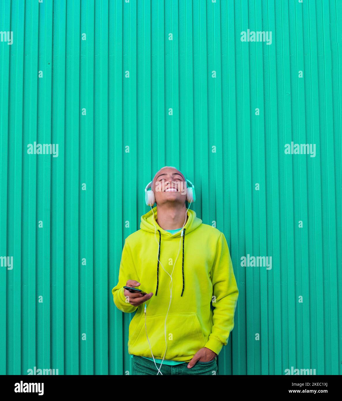 un joven o adolescente milenario escuchando música con su teléfono y auriculares mirando hacia arriba disfrutando de la música y el momento - pelo rubio de hombre de moda - paredes coloridas y suéter - sonriendo y divirtiéndose Foto de stock