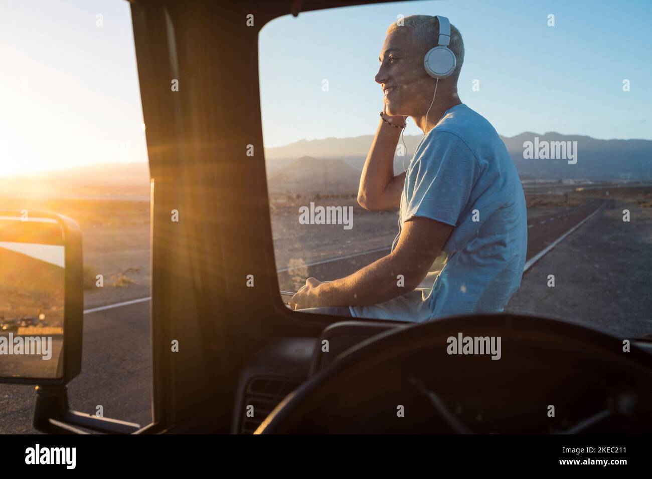 un hombre solo escuchando música con los auriculares en el coche contemplando la hermosa puesta de sol del paisaje, un alegre adolescente viajando y usando su teléfono en sus vacaciones Foto de stock