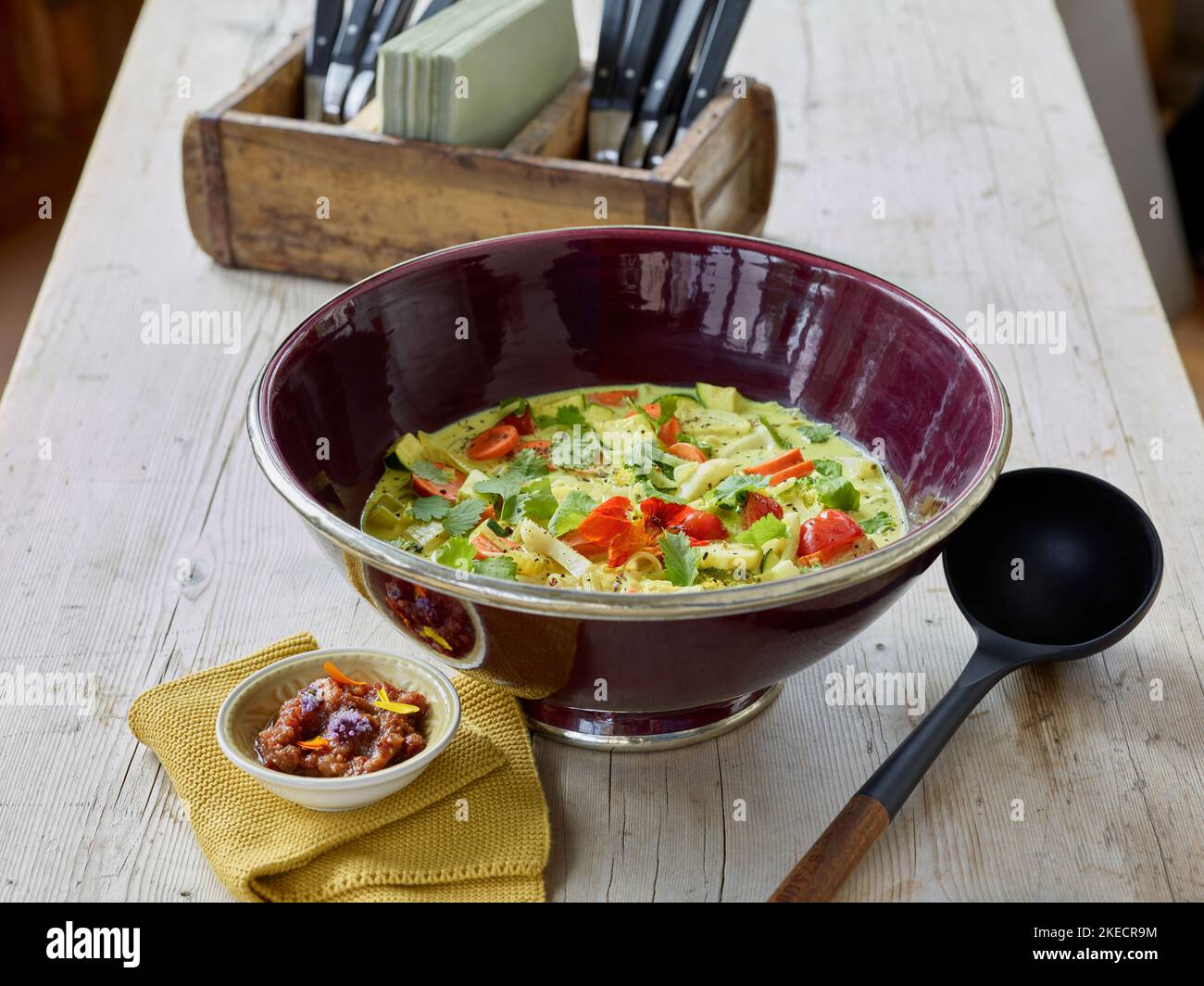 cocina ayurvédica, curry vegetal en un tazón de color berenjena con un tazón pequeño de chutney de pasas de jengibre y cuchara de madera, en el fondo un soporte para cubiertos Foto de stock