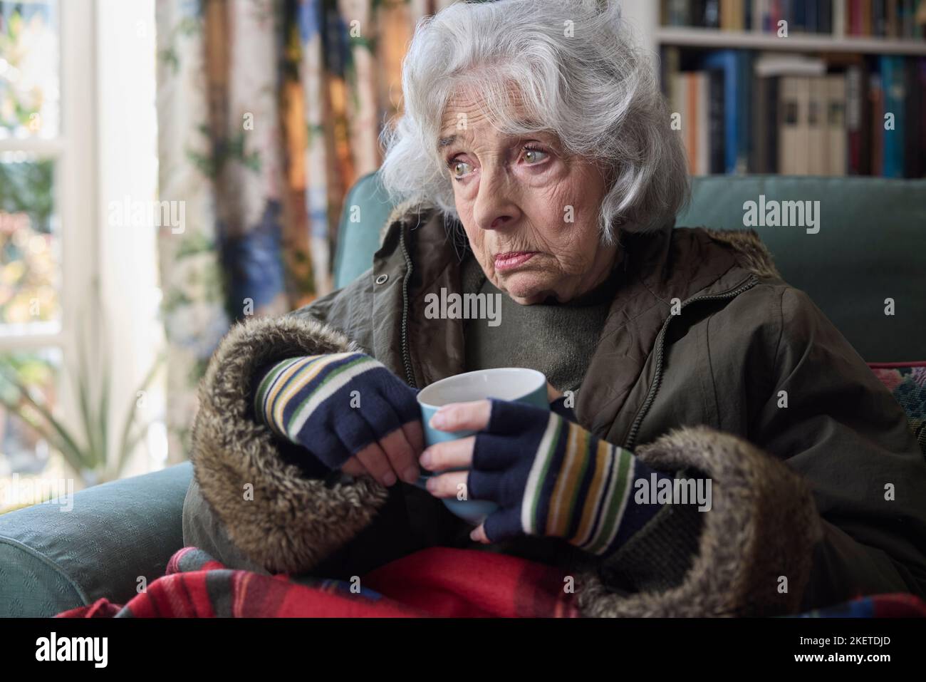 Mujer mayor usando abrigo adentro con bebida caliente que intenta mantener caliente en el hogar en crisis de energía Foto de stock