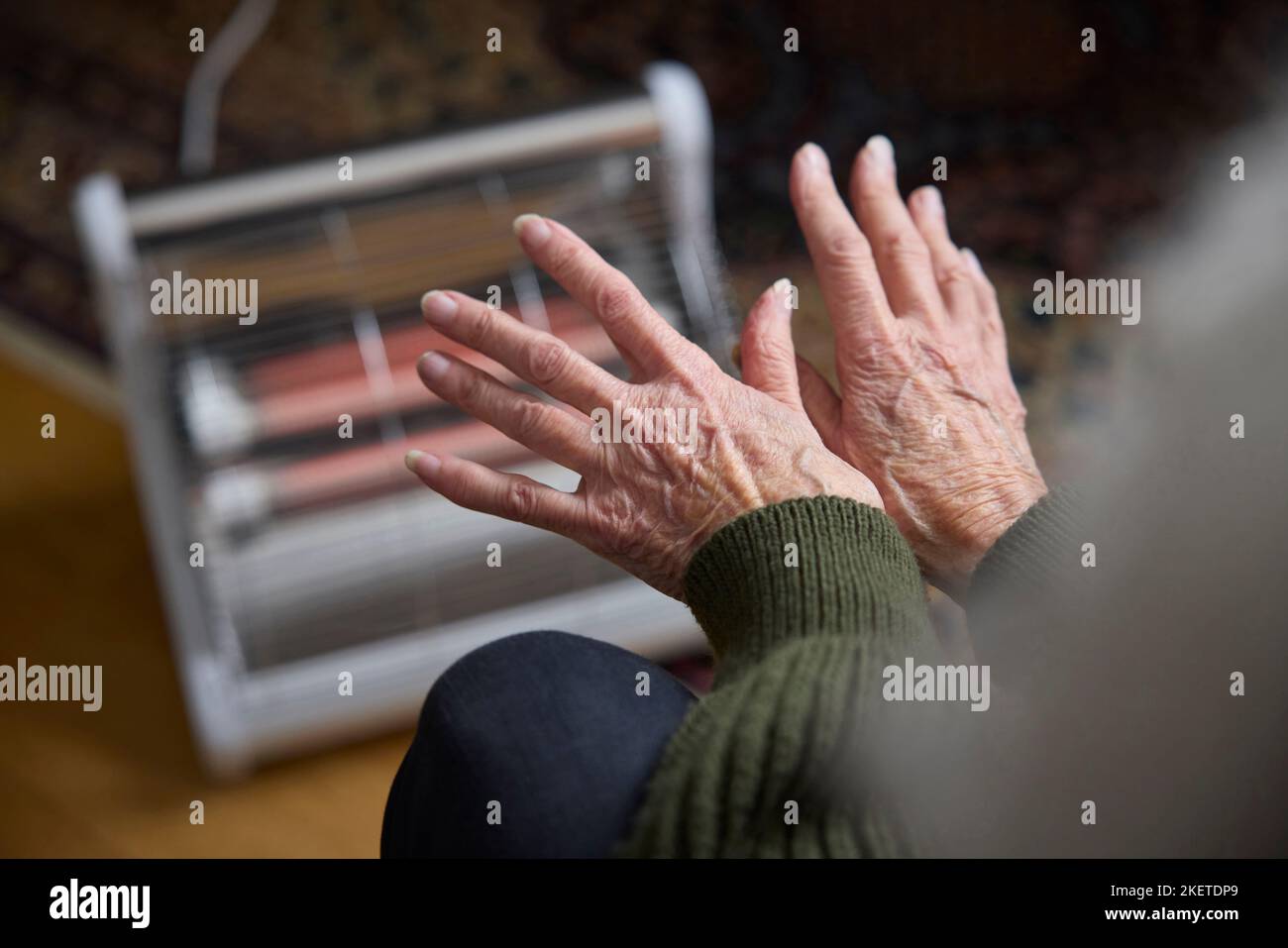 Primer plano de la mujer mayor que calienta las manos en el calentador eléctrico en Cosyt de la crisis de energía viva Foto de stock