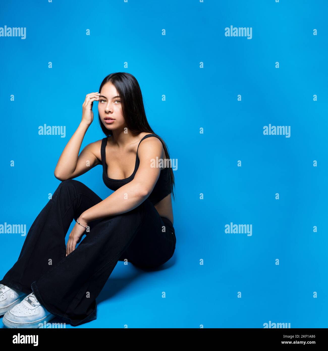 Adolescente multirracial sentado en el piso con las rodillas dobladas rodeado de fondo azul con espacio de copia Foto de stock