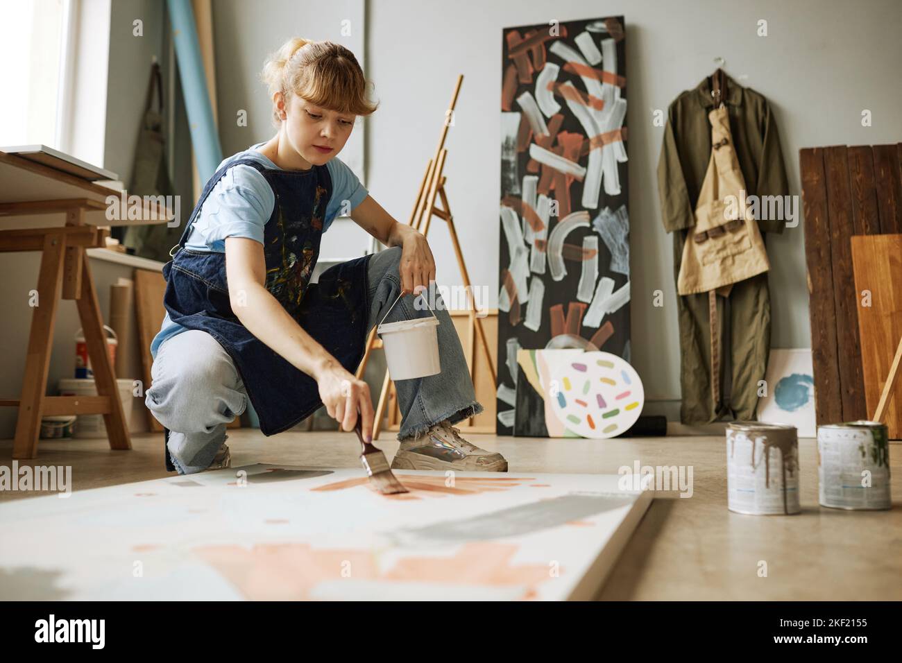 Joven artista rubia con pincel y cubo de plástico pequeño que contiene gouache creando una nueva obra maestra sobre lienzo en estudio Foto de stock