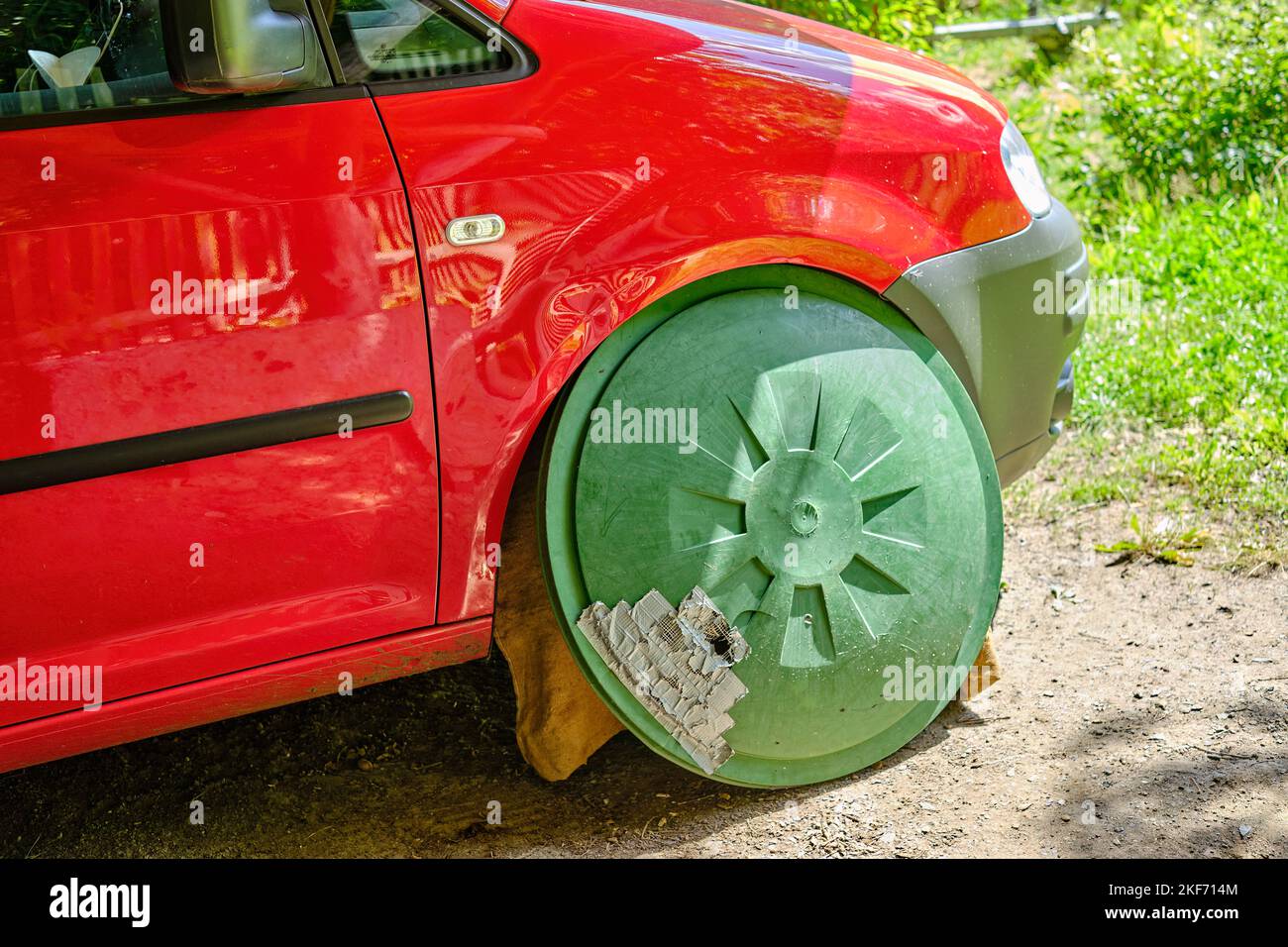 Un coche rojo con una tapa verde de un barril de lluvia donde se supone que la rueda delantera derecha está. Foto de stock