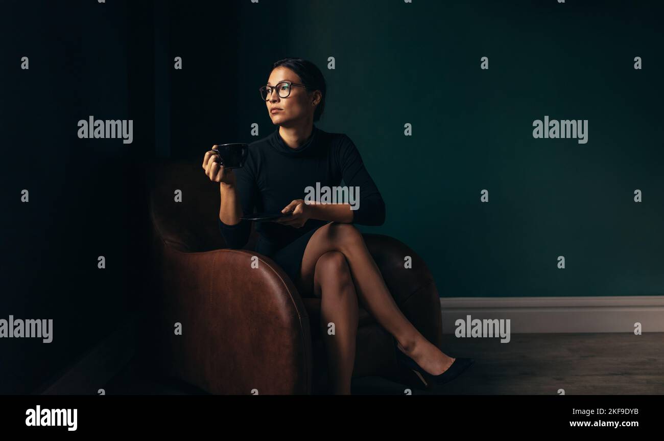 Mujer de negocios joven mirando lejos mientras se sienta en un sofá. Mujer joven de negocios sosteniendo una taza de café en una oficina. Foto de stock