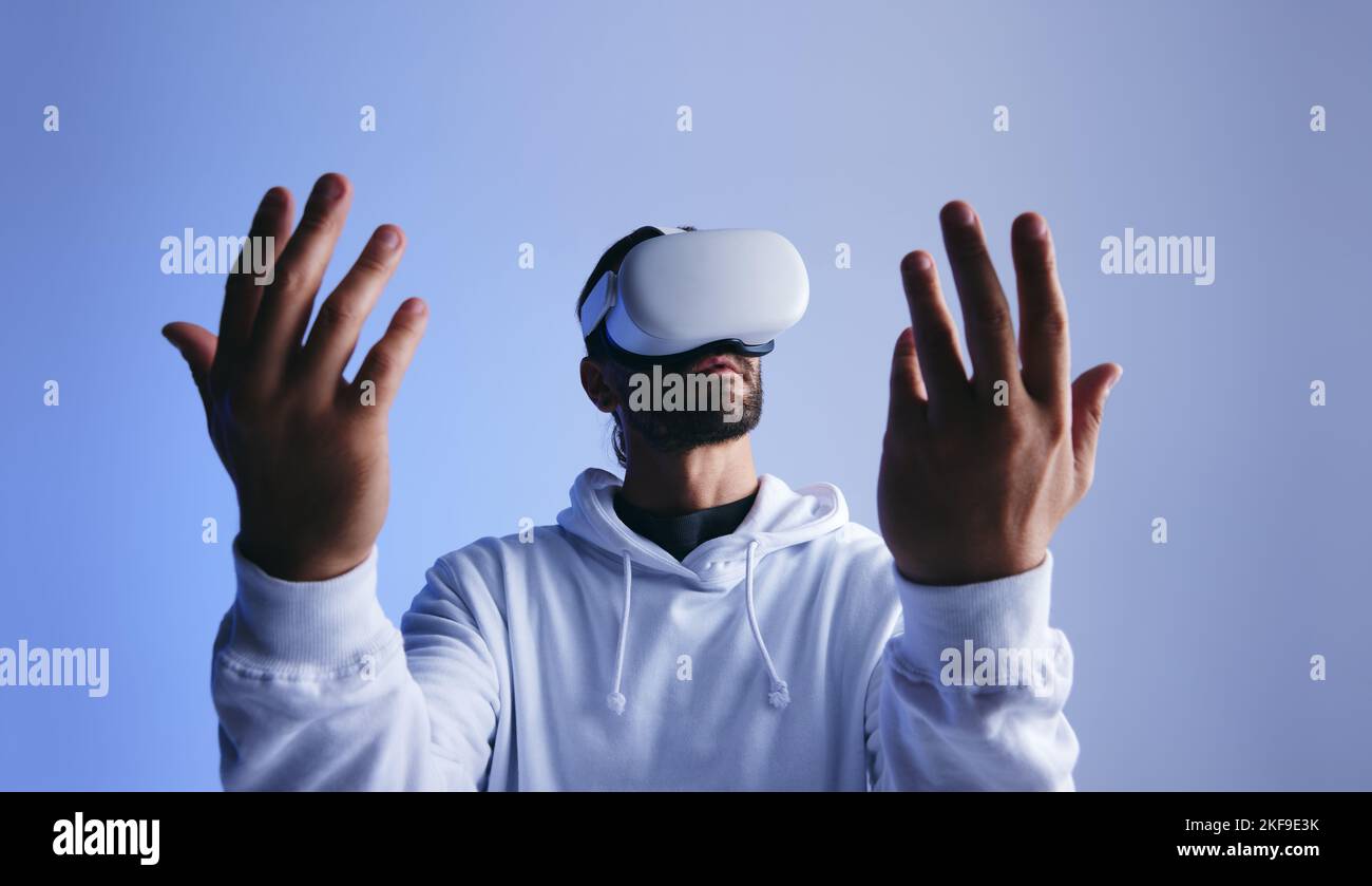 Hombre mirando sus manos a través de gafas de realidad virtual en un estudio. Joven experimentando una simulación en 3D. Joven interactuando con el metaverso Foto de stock