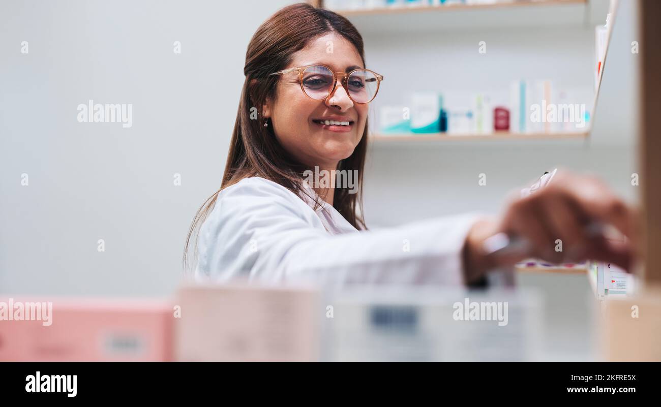 Trabajador de farmacia recibiendo medicamentos de un estante. Farmacéutica india dispensando una receta en un químico. Mujer feliz trabajando en una farmacia. Foto de stock