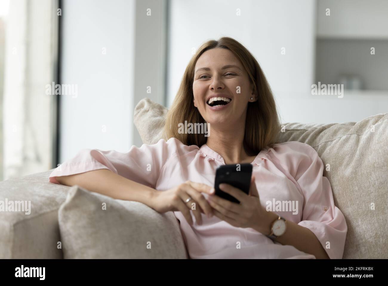 Mujer joven que se ríe Relájese en el sofá con el teléfono móvil Foto de stock