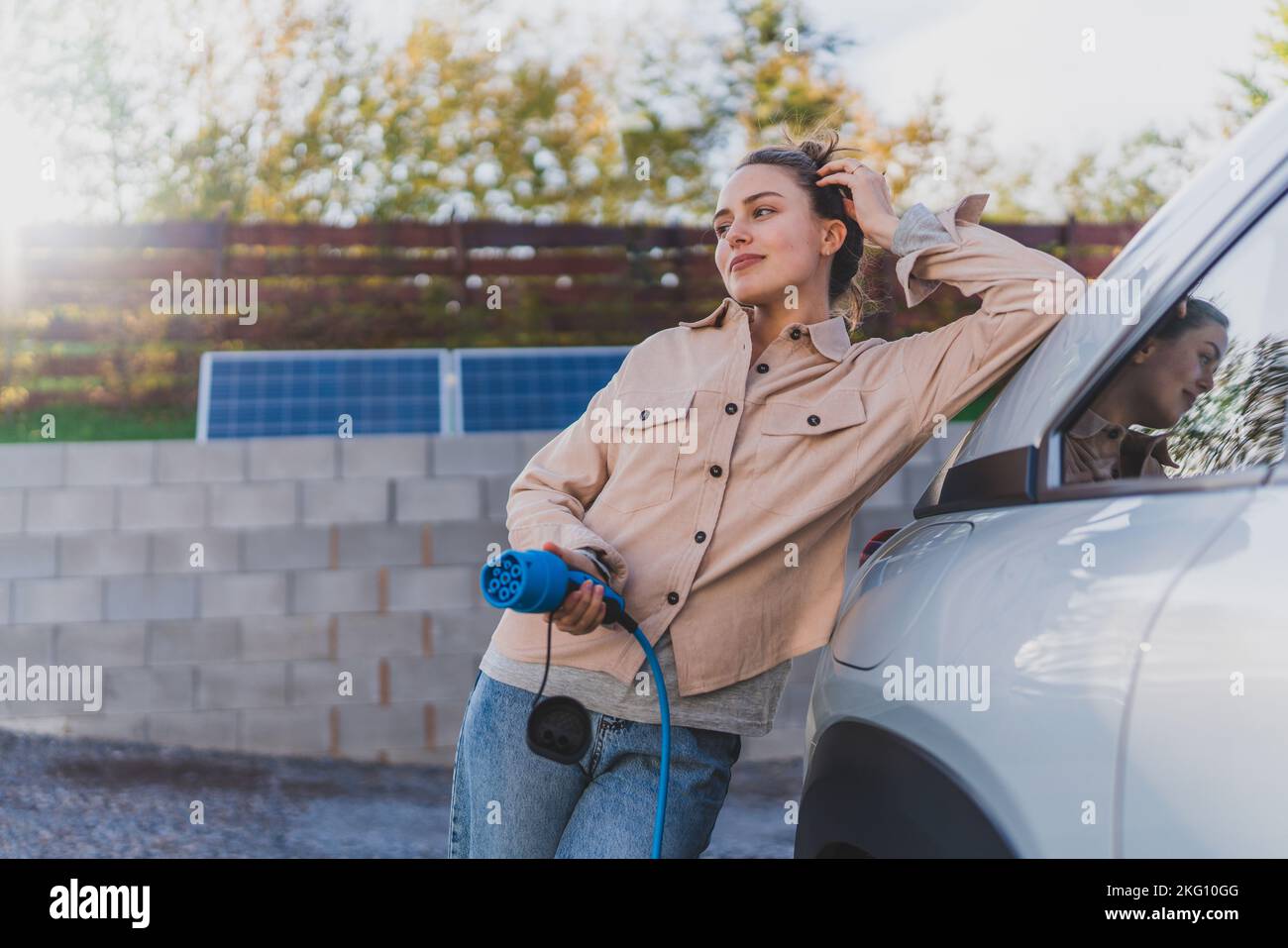 Mujer joven que sostiene el cable de alimentación de su coche eléctrico, preparado para cargarlo en el hogar, sostenible y económico concepto de transporte. Foto de stock