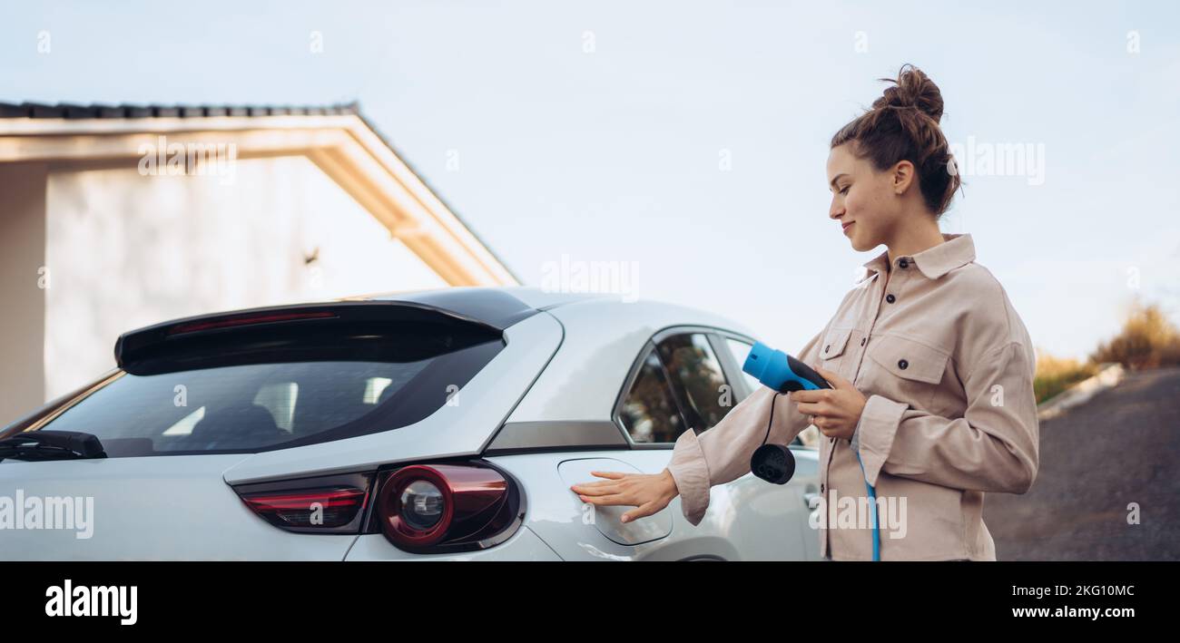 Mujer joven que sostiene el cable de alimentación de su coche eléctrico, preparado para cargarlo en el hogar, sostenible y económico concepto de transporte. Foto de stock