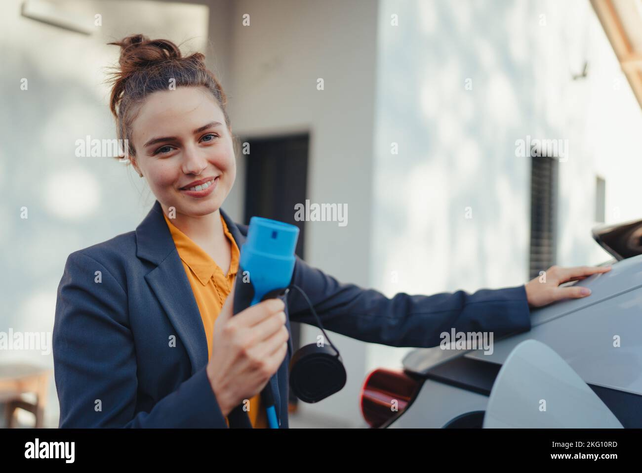 Mujer joven que sostiene el cable de alimentación de su coche eléctrico, preparado para cargarlo en el hogar, sostenible y económico concepto de transporte. Foto de stock