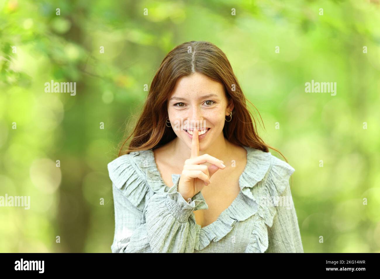 Vista frontal retrato de una mujer feliz pidiendo silencio en un fondo verde del bosque Foto de stock
