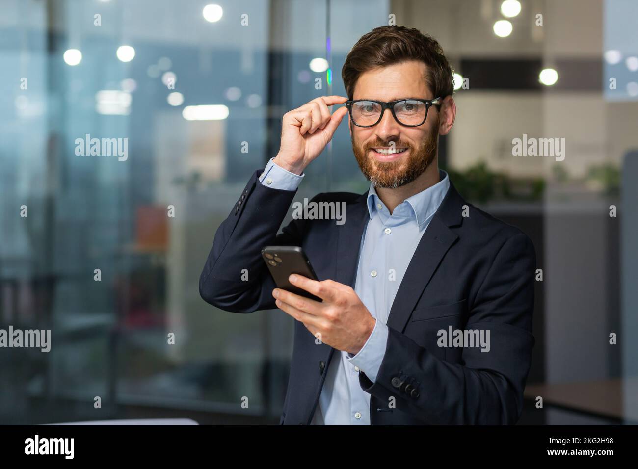 Retrato exitoso jefe con un teléfono, un hombre con gafas y un traje de negocios está mirando la cámara, un inversor está sosteniendo un smartphone en las manos, leyendo un mensaje y utilizando una aplicación en línea. Foto de stock