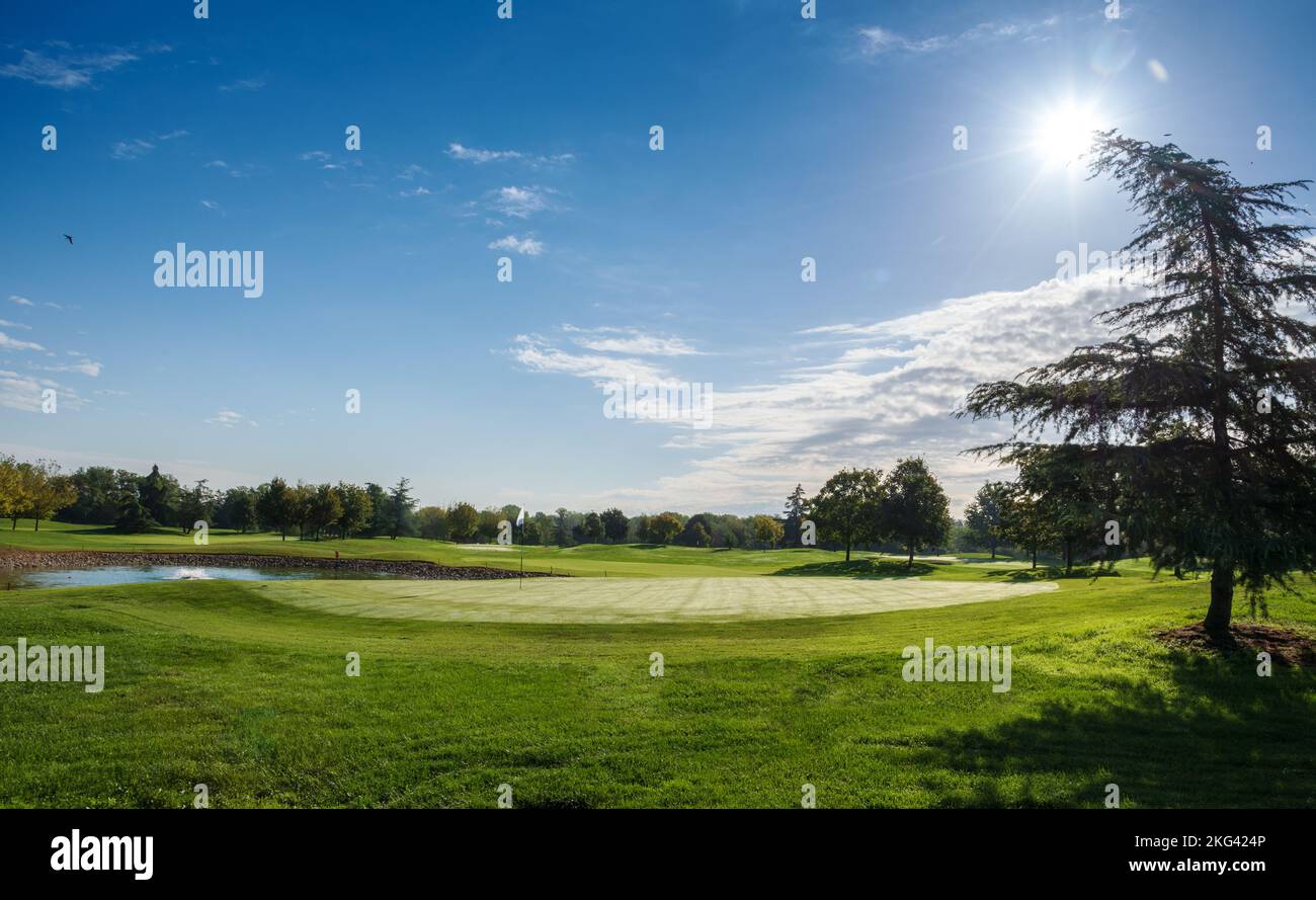Vista panorámica del campo de golf de césped con estanque y árboles situado contra el cielo azul nublado en un soleado día de verano Foto de stock