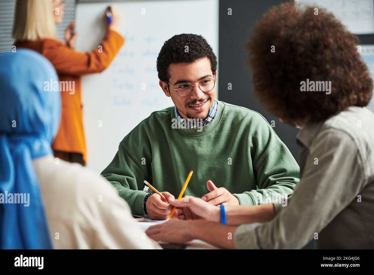 Hombre joven feliz en anteojos comunicándose con otro estudiante en el descanso entre las lecciones contra el maestro preparándose para la presentación Foto de stock