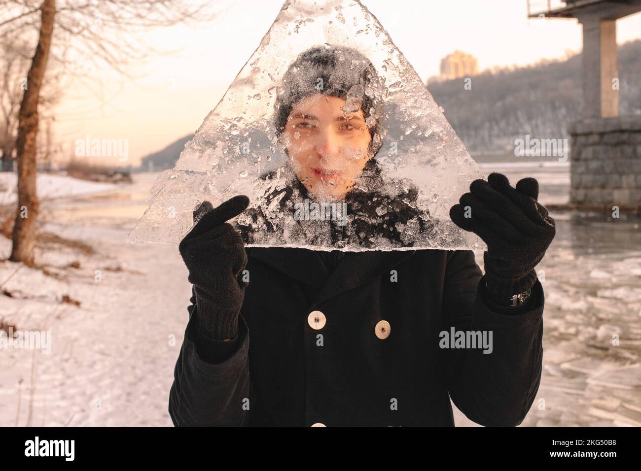 Adolescente sosteniendo un pedazo de hielo delante de su cara y mirando a través del hielo mientras estaba de pie al aire libre en clima frío Foto de stock