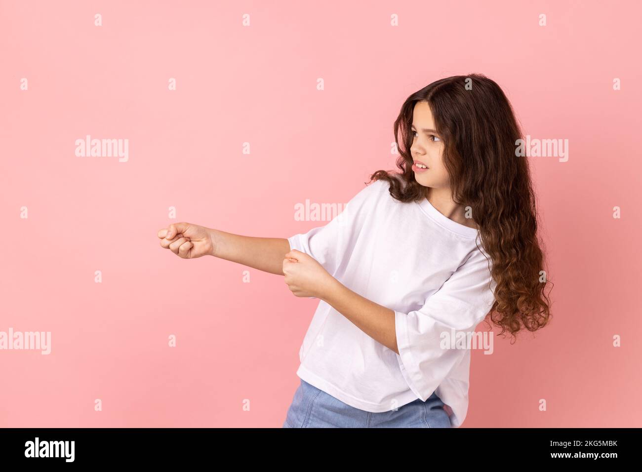 Vista lateral retrato de encantadora niña adorable con camiseta blanca de pie en ataque o tirando de las manos gesto y mirando hacia adelante. Estudio de interior grabado aislado sobre fondo rosa. Foto de stock