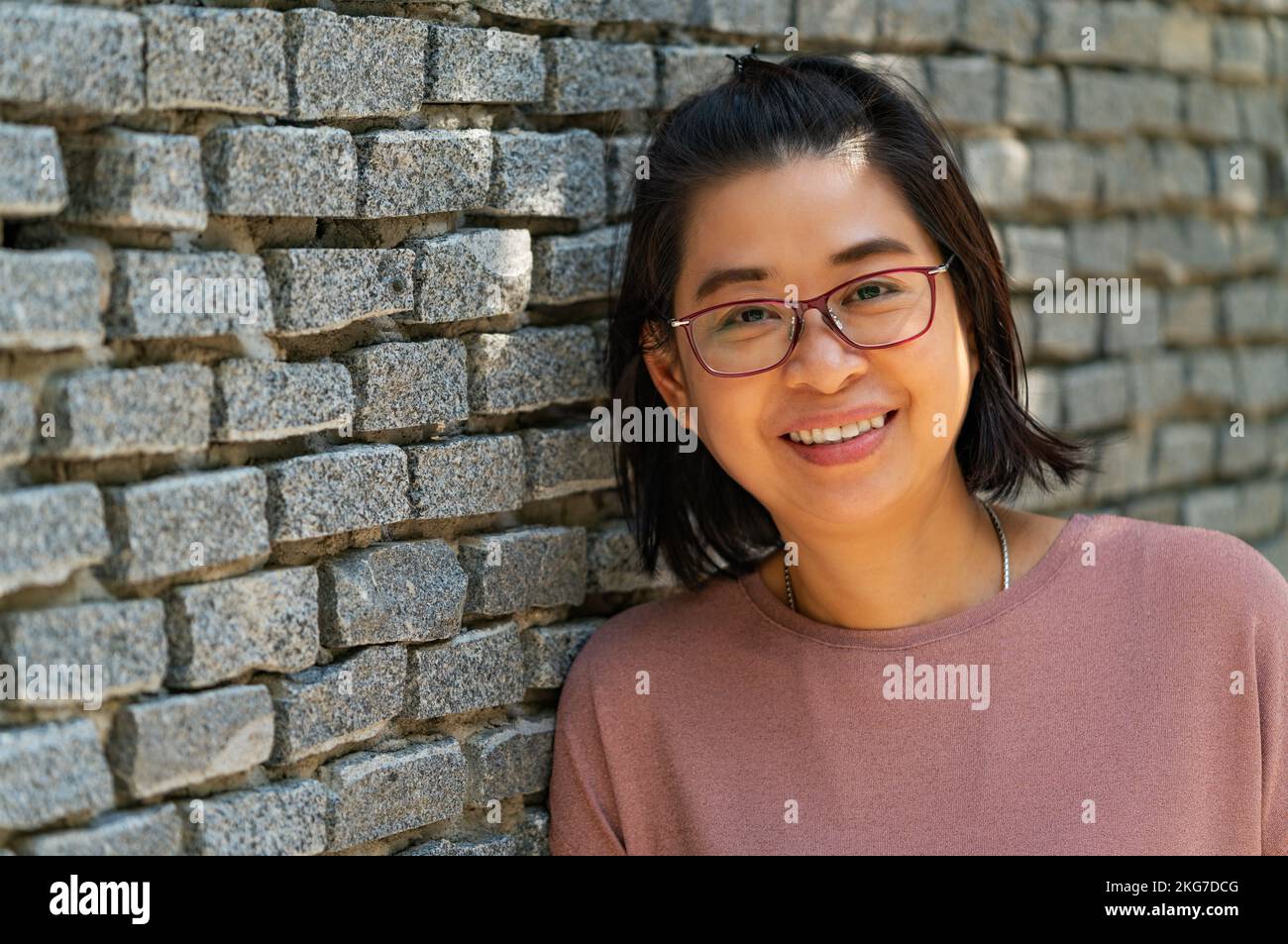 Retrato mujer asiática de mediana edad con rostro sonriente, sonriendo, se ve saludable, usa gafas, se apoya en pared gris de ladrillo, vista en perspectiva, s vacío Foto de stock