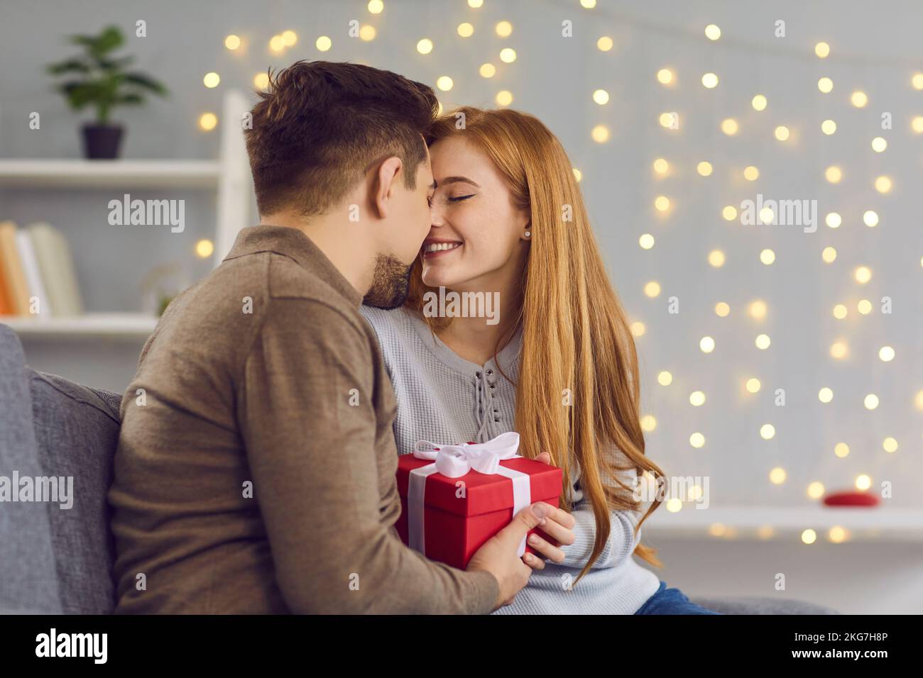 Feliz pareja en el amor sentado en el sofá en casa, sosteniendo el presente y tocando las narices a punto de besarse Foto de stock