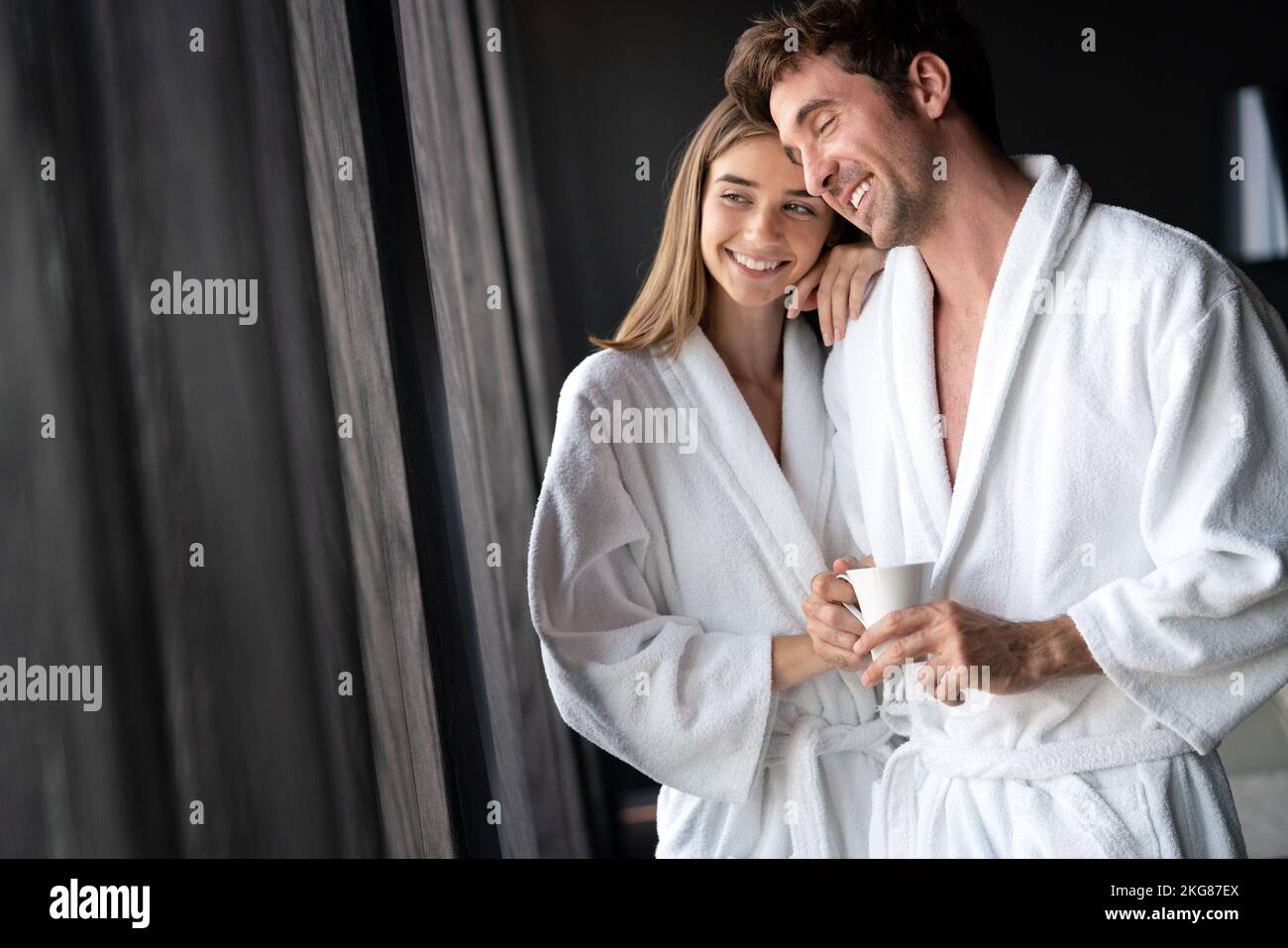 Feliz pareja joven en batas blancas bebiendo café juntos. Hotel, viajes, relaciones concepto Foto de stock