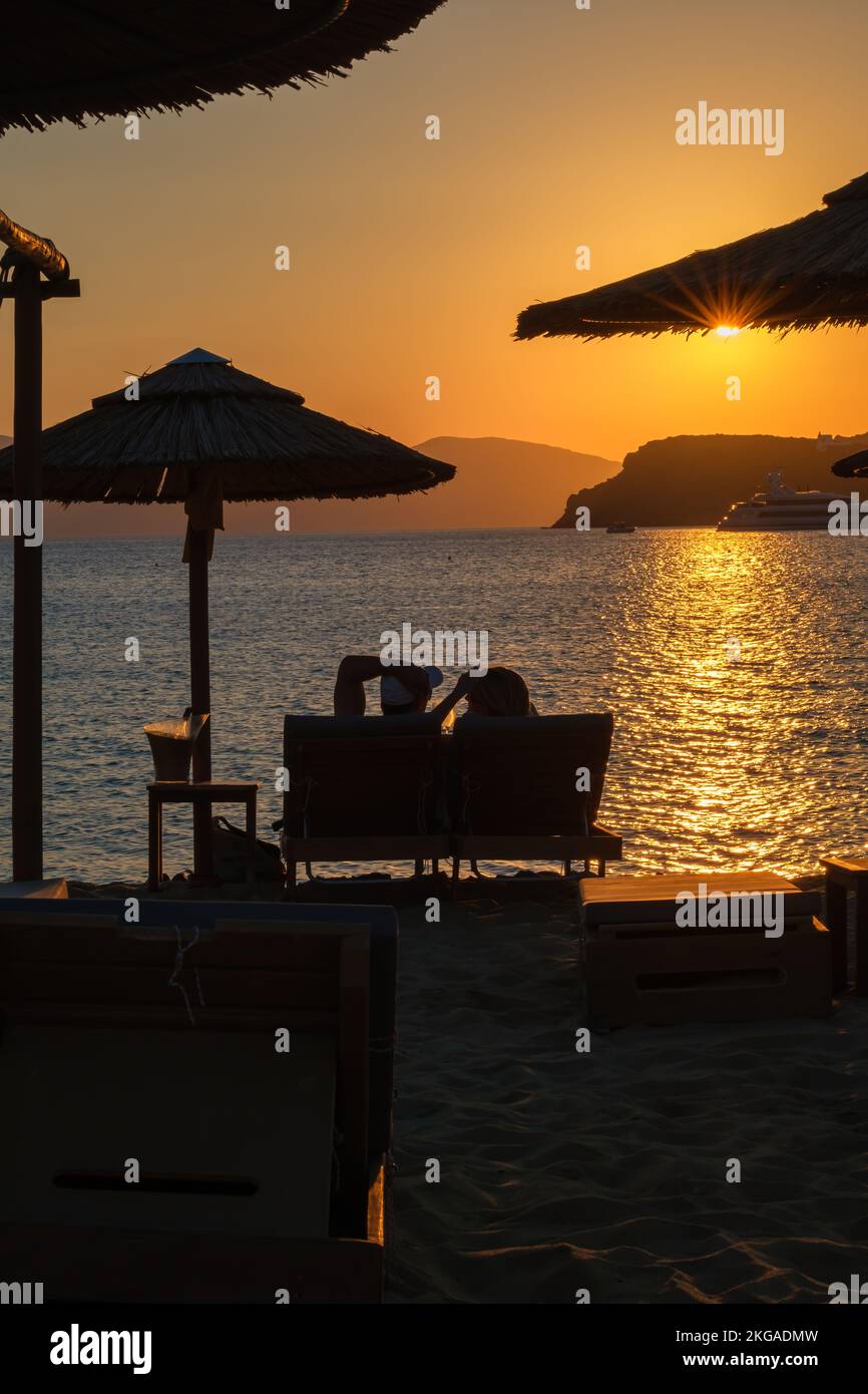 IOS, Grecia - 13 de septiembre de 2022 : Vista de una pareja acostada en las tumbonas, bebiendo vino y disfrutando de la increíble puesta de sol naranja en Ios Grecia Foto de stock