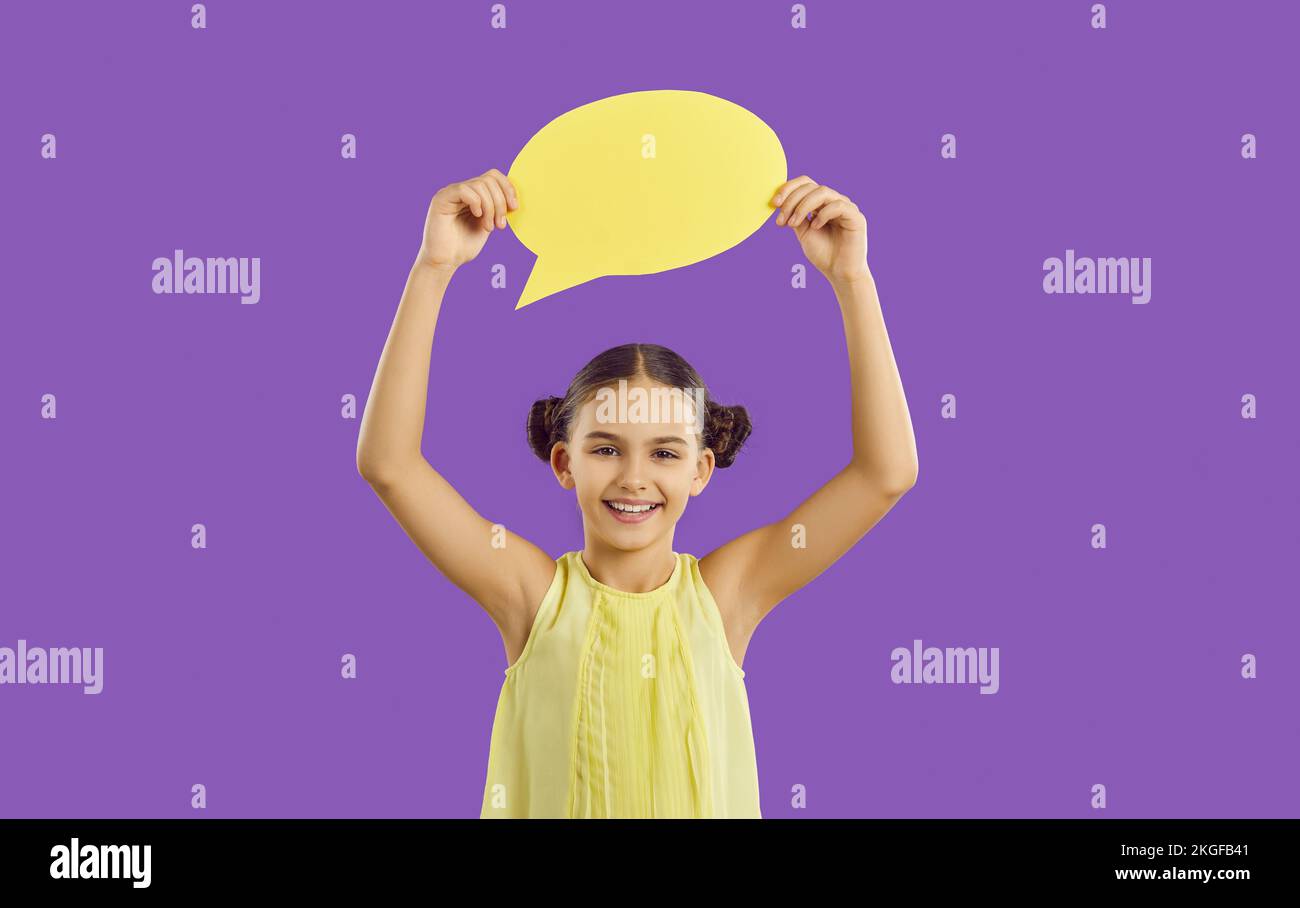 Chica feliz de pie sobre fondo púrpura, mostrando burbuja de habla amarilla y sonriendo Foto de stock