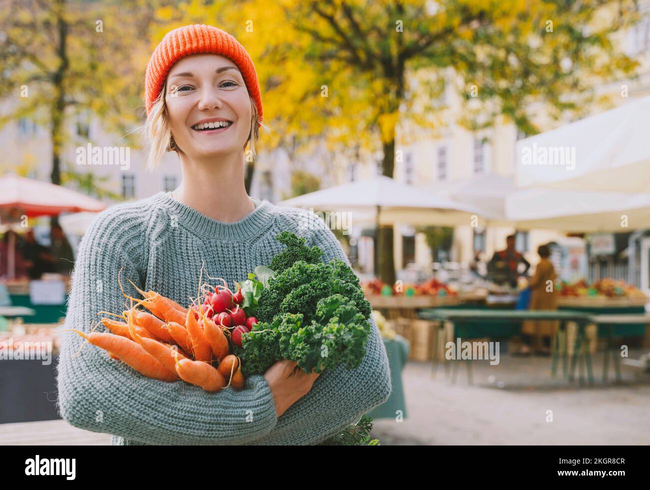 Mujer feliz usando suéter sosteniendo verduras orgánicas en el mercado Foto de stock