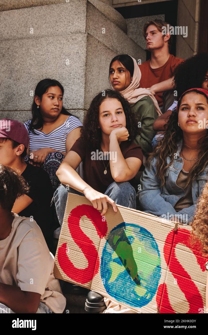 Activistas juveniles multiculturales sentados fuera de un edificio con un estandarte de 'guardar nuestra especie'. Grupo de jóvenes diversos que protestan contra la guerra mundial Foto de stock