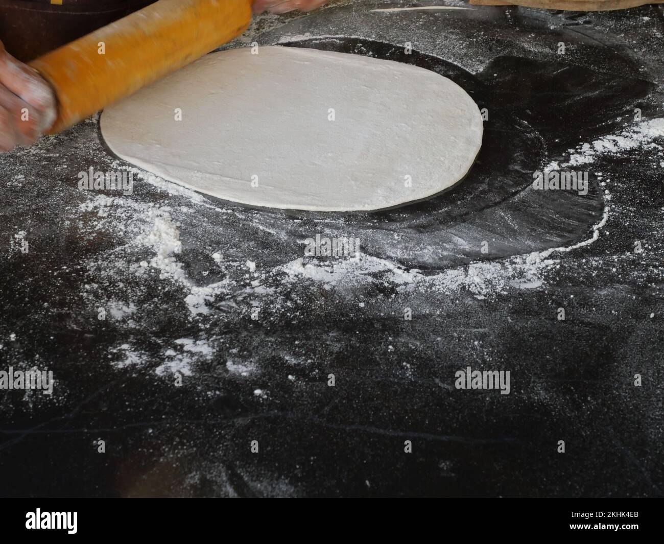 La mano del chef está rodando masa de pizza en forma de círculo aplanado por un rodillo de madera con harina sobre fondo negro Foto de stock