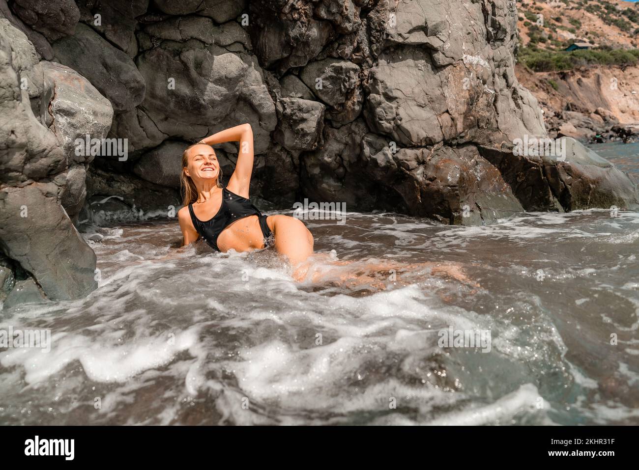 Mujer traje de baño mar. Atractiva mujer rubia en un traje de baño negro disfrutando del aire del mar en la orilla alrededor de las rocas. Concepto de viajes y vacaciones. Foto de stock
