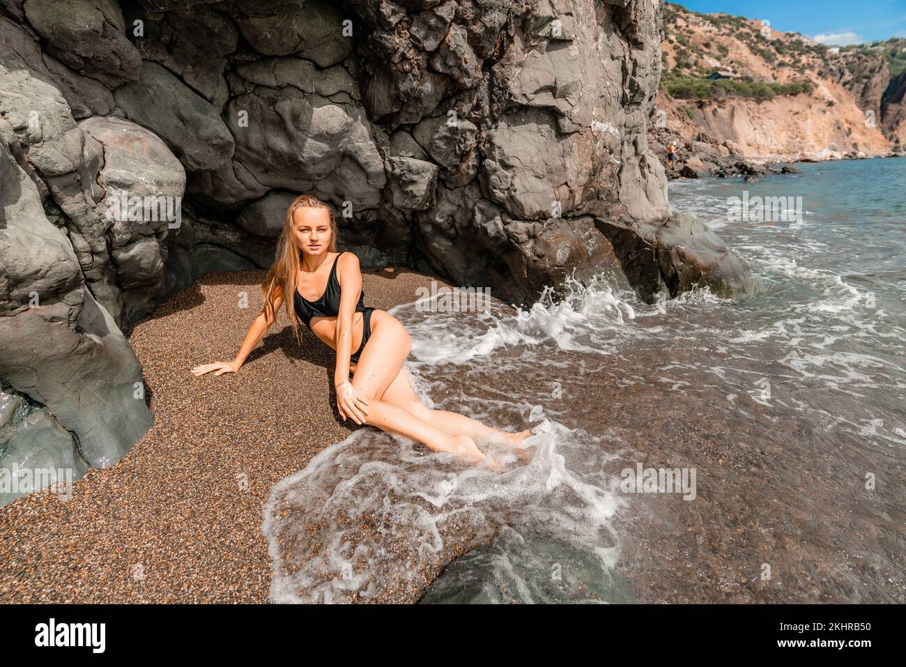 Mujer traje de baño mar. Atractiva mujer rubia en un traje de baño negro disfrutando del aire del mar en la orilla alrededor de las rocas. Concepto de viajes y vacaciones. Foto de stock