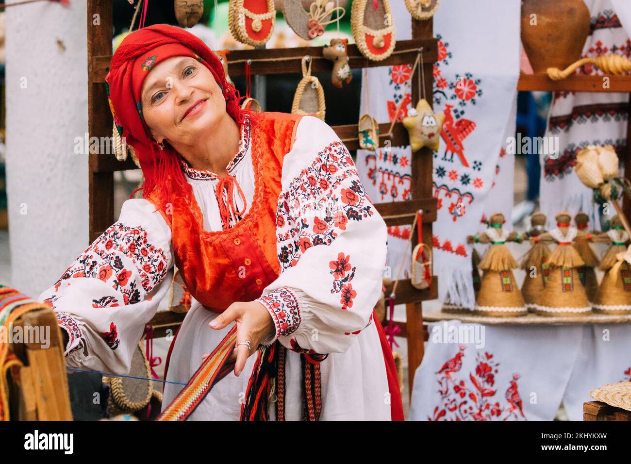 Tejido femenino para adultos sonriente en ropa tradicional bordada bielorrusa muestra el proceso de hilado de cinturón. Festival Folk en Minsk, Bielorrusia. Foto de stock