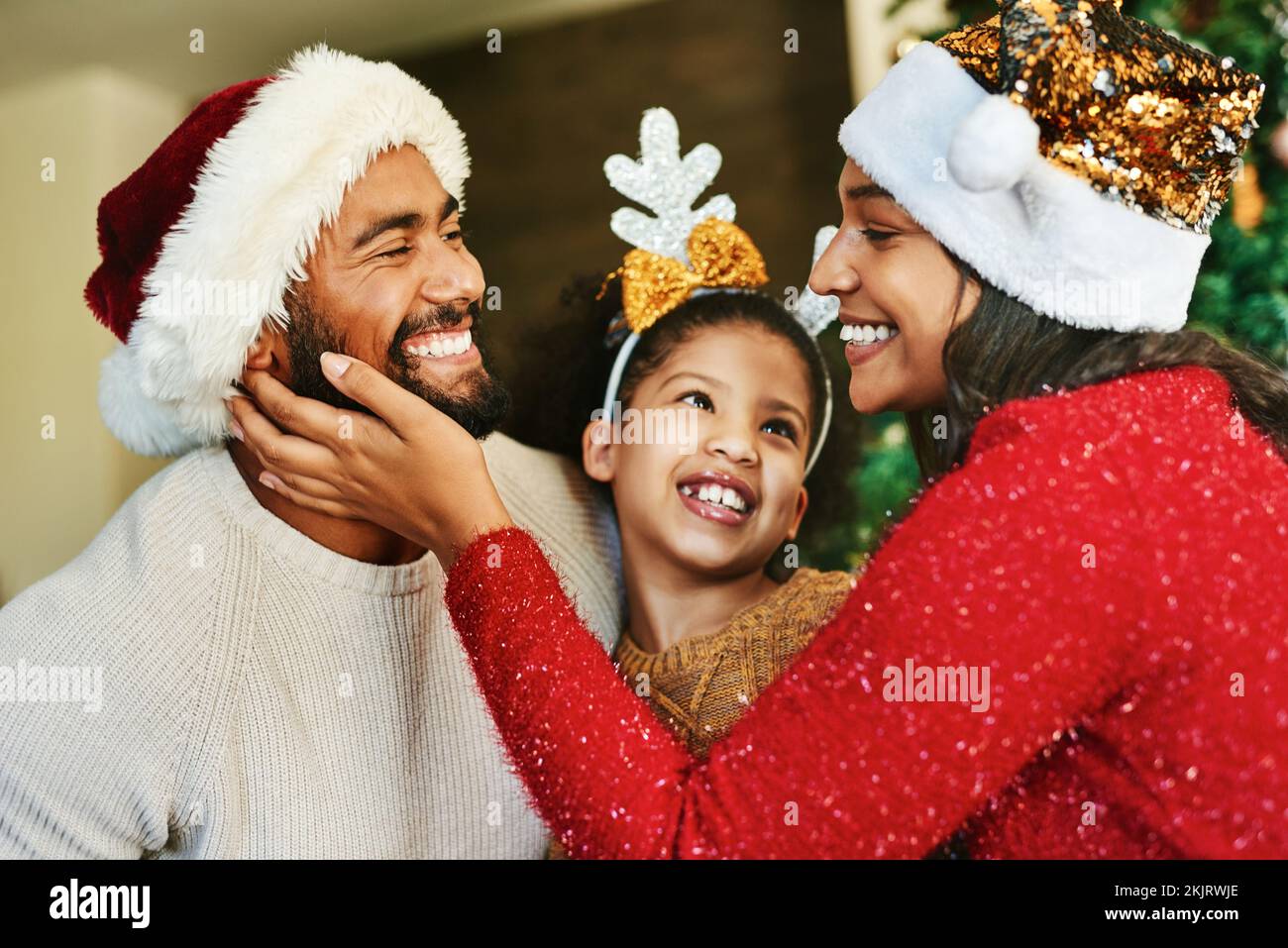 Familia, celebración de navidad y amor con una sonrisa feliz y vacaciones festivas, cuidado. y decoración con sombrero en casa. Madre, padre y niña juntos Foto de stock