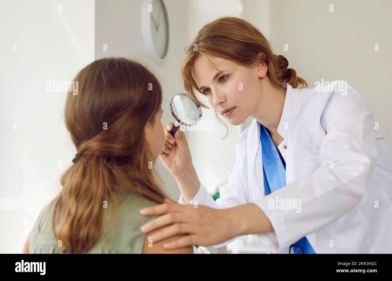 Dermatóloga profesional examina la cara de una adolescente con una lupa en su oficina. Foto de stock