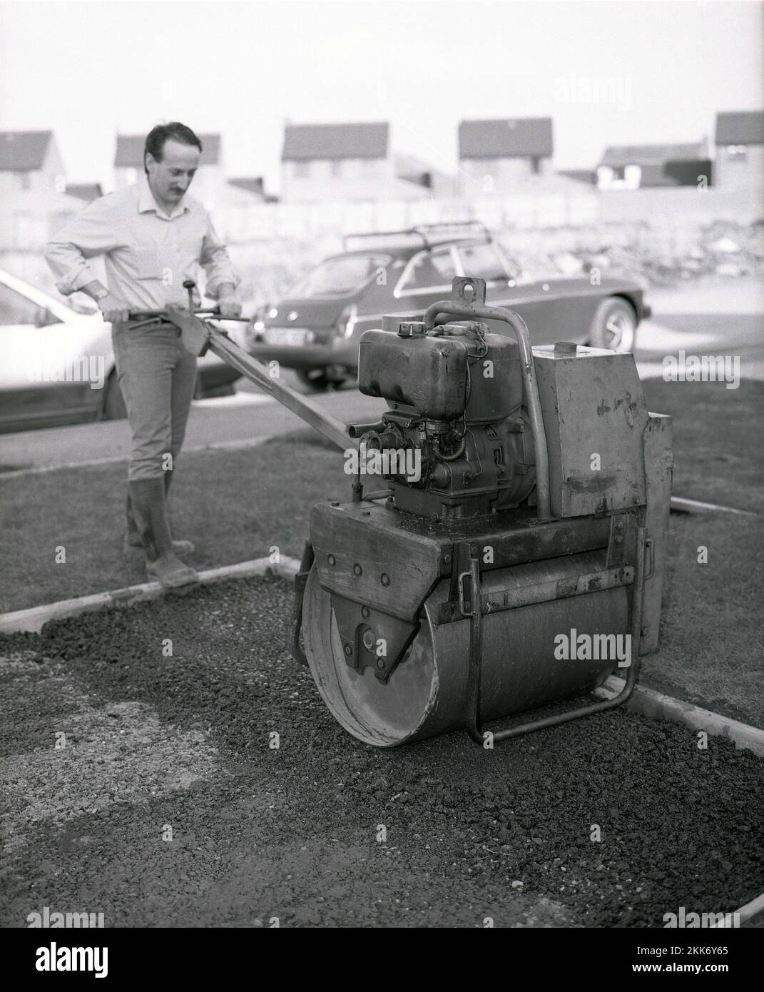 1980s, histórico, un hombre repavimentando un camino de una casa, usando un pequeño rodillo de la carretera de la época, también conocido como compactador, para acostarse en el asfalto, Inglaterra, Reino Unido. Foto de stock