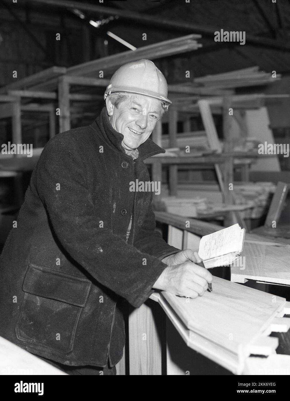 1980s, histórico, en el interior de un almacén de materiales de construcción de madera, un hombre casa constructor en una chaqueta burro y casco, con cuaderno, con medidas, Inglaterra, Reino Unido Foto de stock