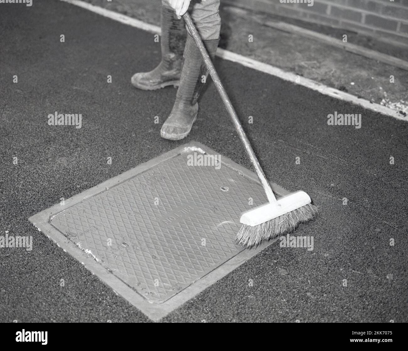 1980s, re-surfacing una calzada, un trabajador usando una escoba para cepillar cualquier residuo negro de macadam dejado en una tapa de la boca de inspección después de asfaltar una calzada de la casa, Inglaterra, Reino Unido. Foto de stock