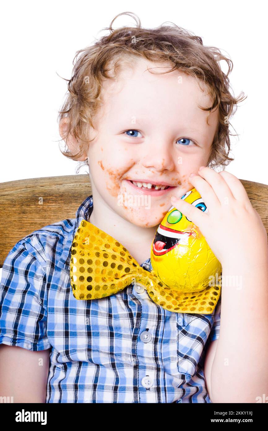 Niño caucásico con ojos azules y cabello rubio rizado sucio con camisa azul cuadros y lazo amarillo brillante. Él sostiene un huevo amarillo y tiene una choc Foto de stock
