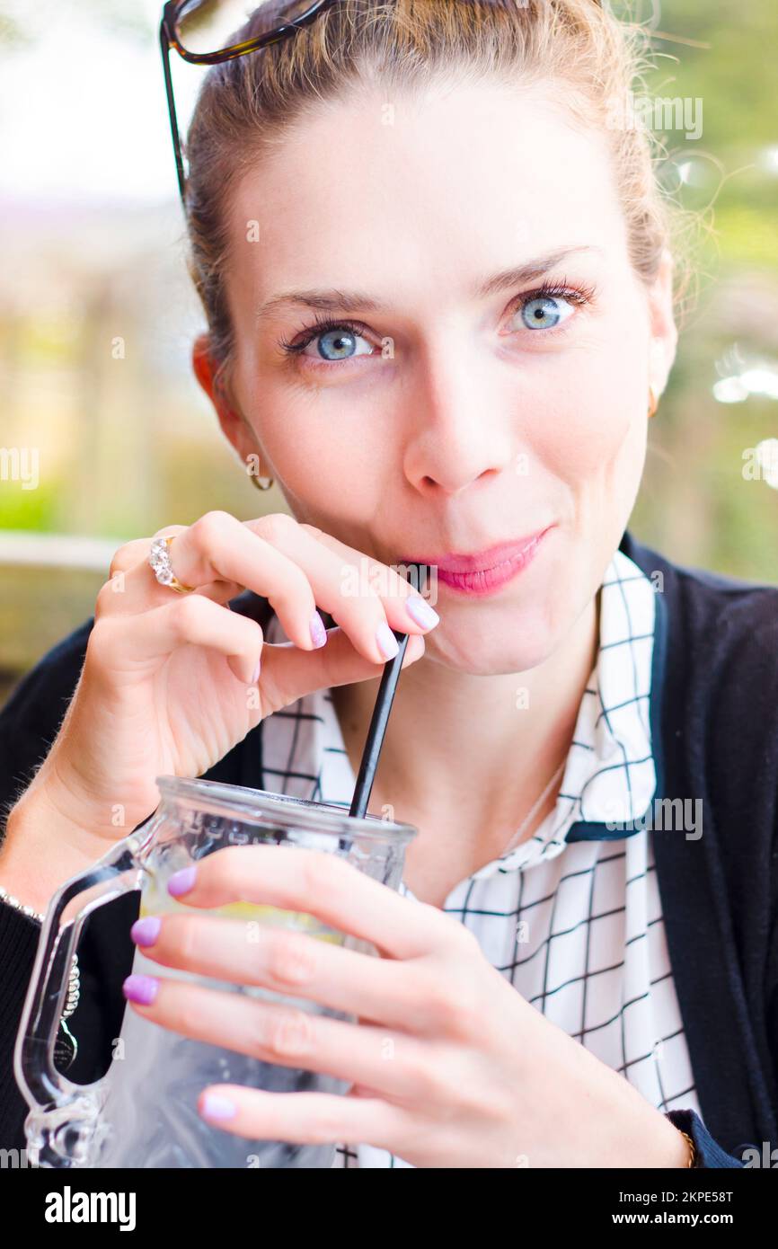 Hermosa mujer joven a principios de los años veinte mirando la cámara con sonríe al beber un vaso frío de agua con un trozo de lemon en un restaurante al aire libre Foto de stock