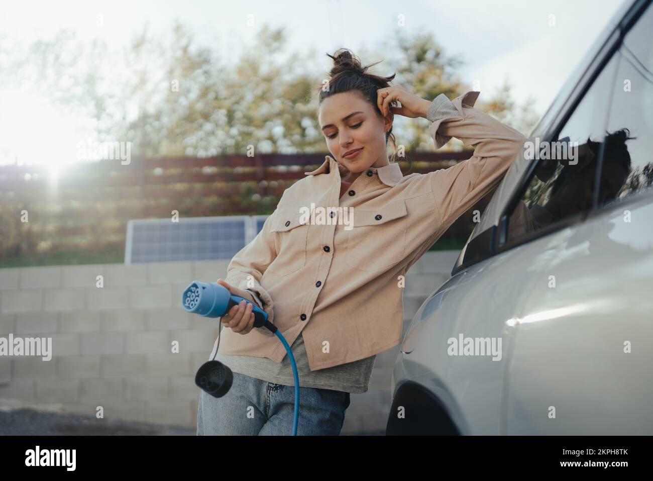 Mujer joven que sostiene el cable de alimentación de su coche eléctrico, preparado para cargarlo en el hogar, sostenible y económico concepto de transporte. Foto de stock