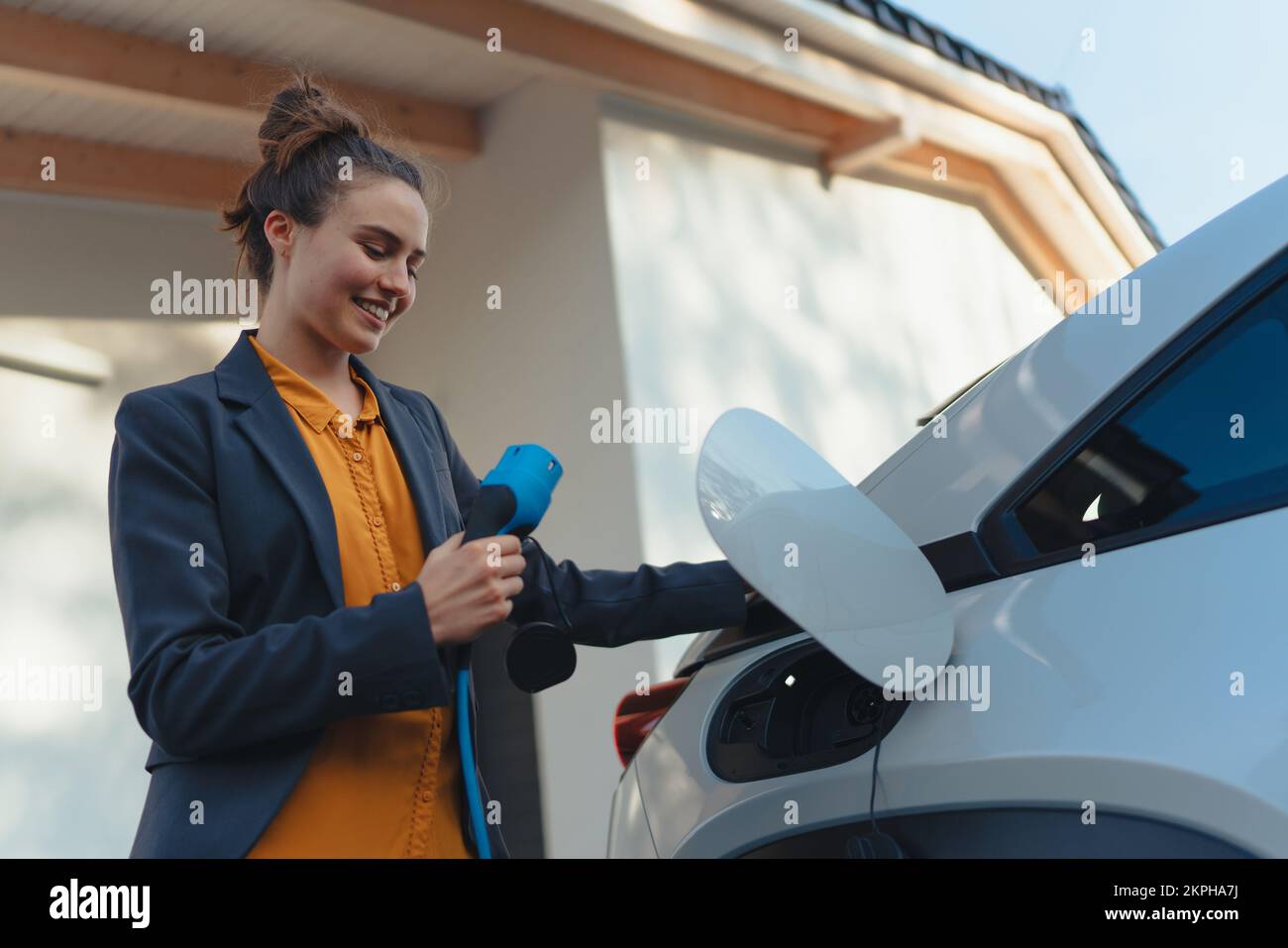 Mujer joven que sostiene el cable de alimentación de su coche eléctrico, preparado para cargarlo en el hogar, sostenible y económico concepto de transporte. Foto de stock