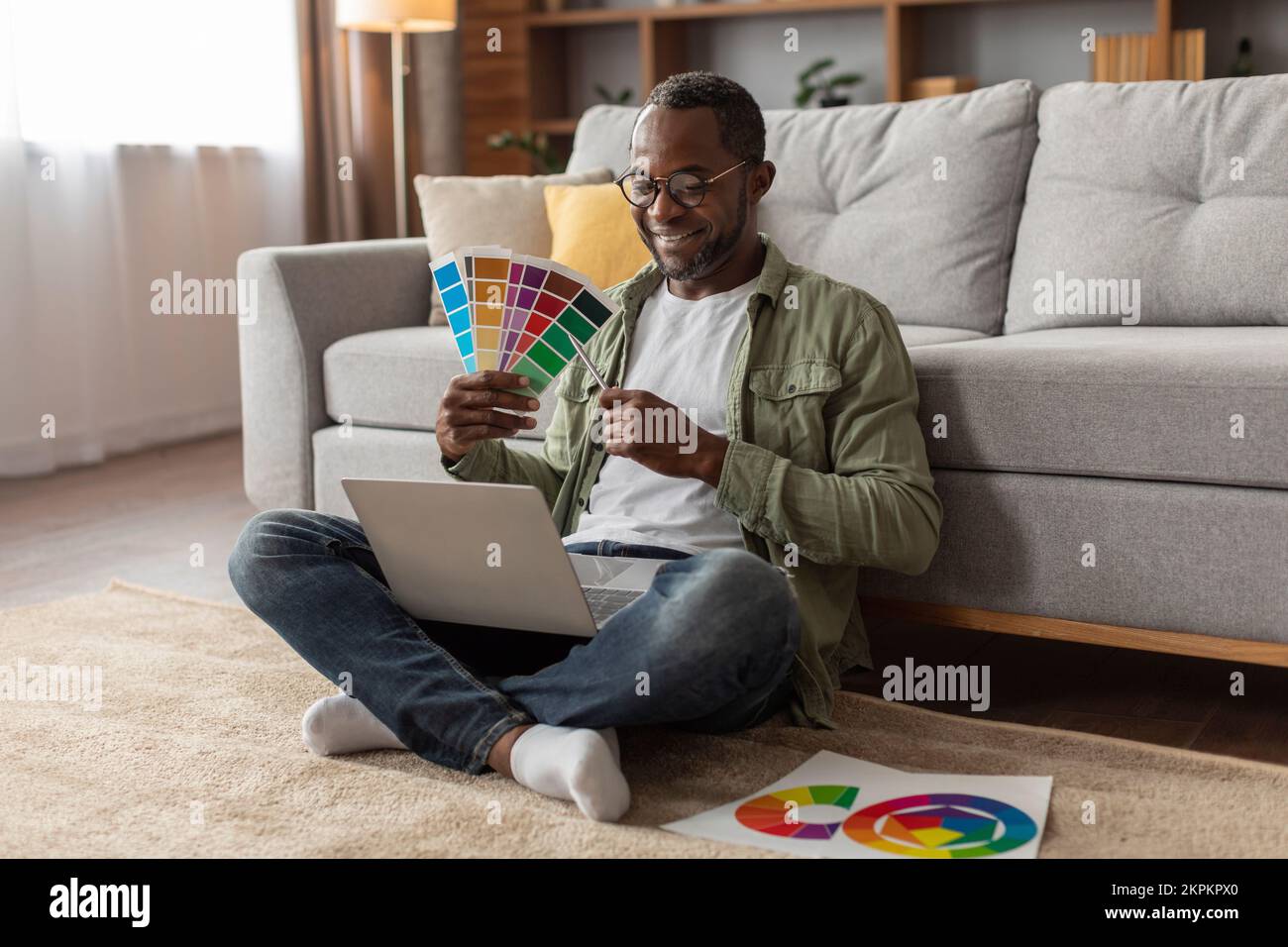 Sonriente diseñador afroamericano de mediana edad en gafas muestra paletas de color en webcam portátil Foto de stock
