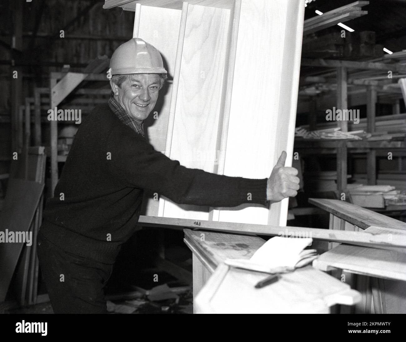 1980s, histórico, un capataz de la construcción en un casco dentro de un almacén del edificio que sostiene una pequeña escalera de madera, para ser puesto en una nueva casa, Inglaterra, Reino Unido. Foto de stock