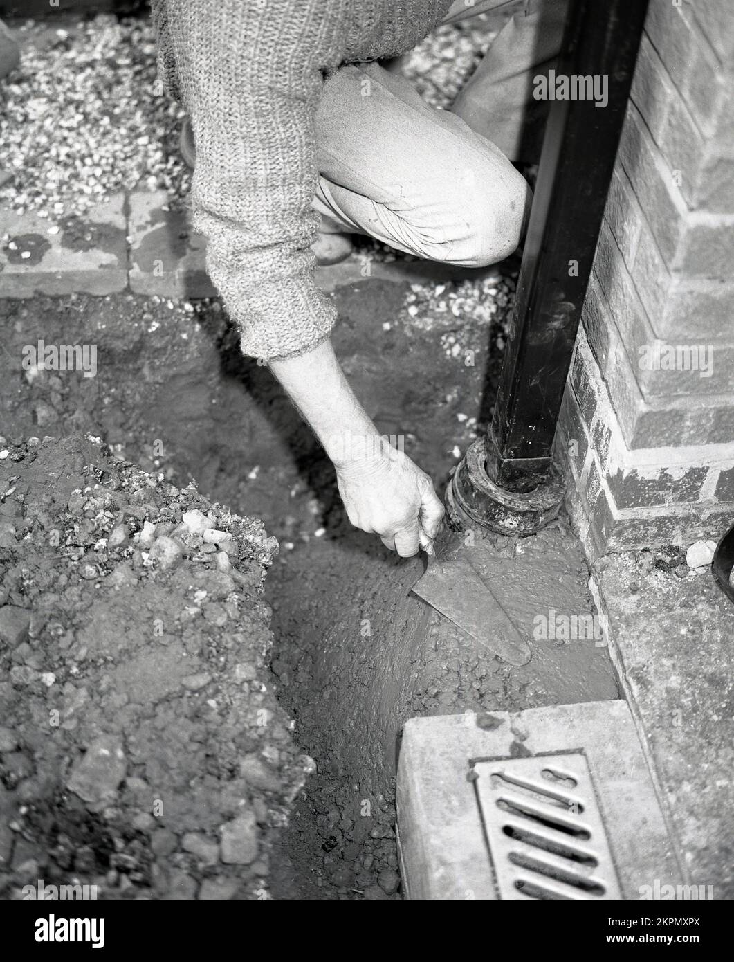 1980s, un trabajador que usa una mezcla lista o listo poner tarmacadam frío para volver a pavimentar una pequeña área por un desagüe cubierto de ladrillo en un camino de entrada en el frente de una casa, Inglaterra, Reino Unido, Foto de stock