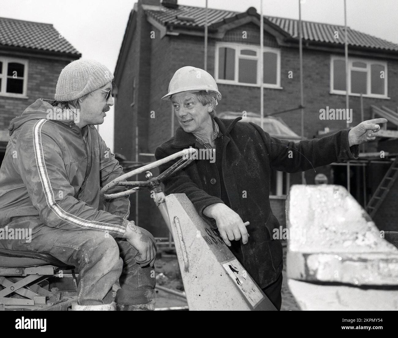1980s, fuera en un sitio de construcción de una nueva vivienda, un capataz usando casco, dando instrucciones a un trabajador de la construcción usando un sombrero de bolitas y anorak, que está en un dumper de punta, un vehículo de construcción que es un tipo de camión de dumper, Inglaterra, Reino Unido. Foto de stock