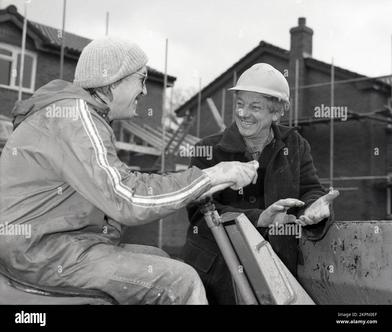 1980s, dos constructores en obra, teniendo una conversación y una risa, uno usando un casco, el otro un sombrero de bolitas, que está sentado en un dumper de la punta, un vehículo de la construcción que es un tipo de carro del dumper, Inglaterra, Reino Unido. Foto de stock