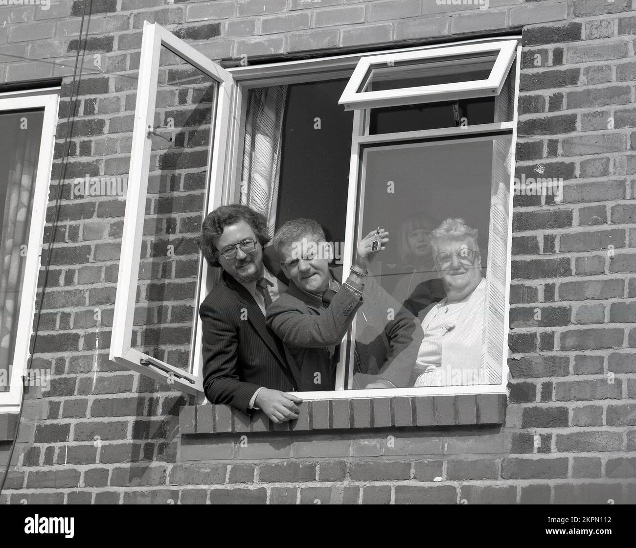 1980s, histórico, de pie en una ventana de su nuevo hogar con un ejecutivo de vivienda una pareja con su hija posan para una foto de publicidad, con el hombre sosteniendo las llaves, Inglaterra, Reino Unido. Foto de stock