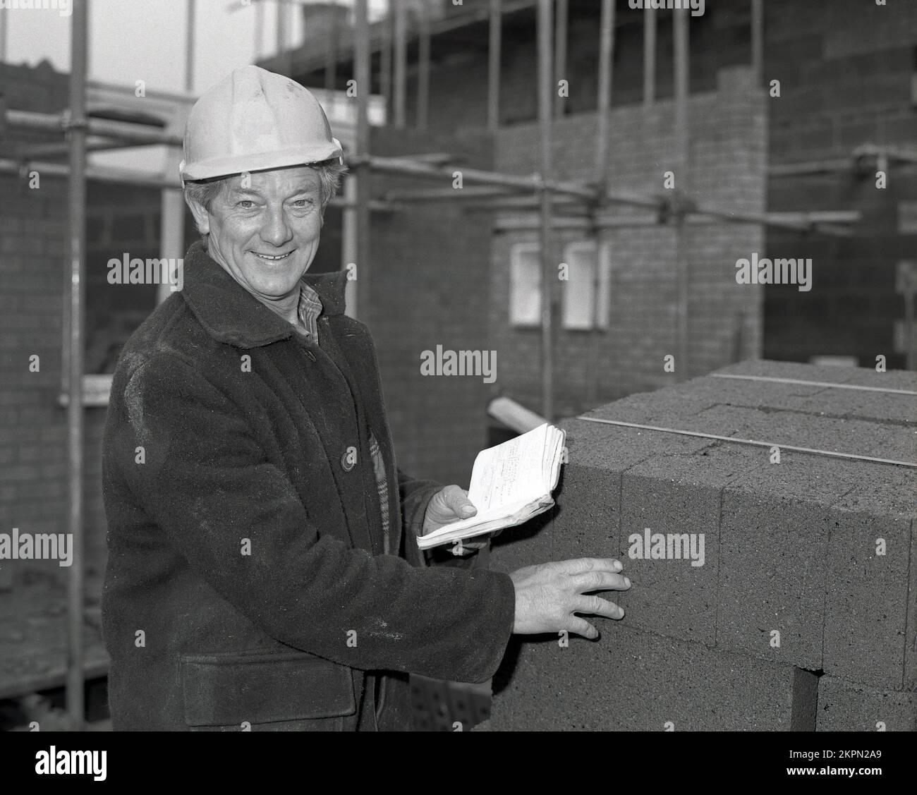 1980s, en un sitio de construcción de viviendas, un capataz en una chaqueta de burro y casco sujetando un cuaderno, mientras comprueba una pila de bloques de hormigón ligero, utilizados en la construcción de nuevas viviendas, Inglaterra, Reino Unido. Foto de stock