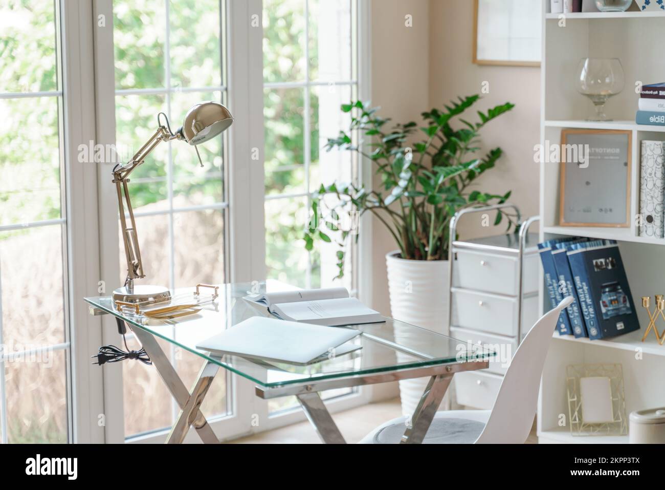 Lugar de trabajo cómodo con un ordenador portátil en la oficina y en casa. Habitación luminosa con grandes ventanas, estantería y planta verde. Trabajo remoto, independiente. Foto de stock